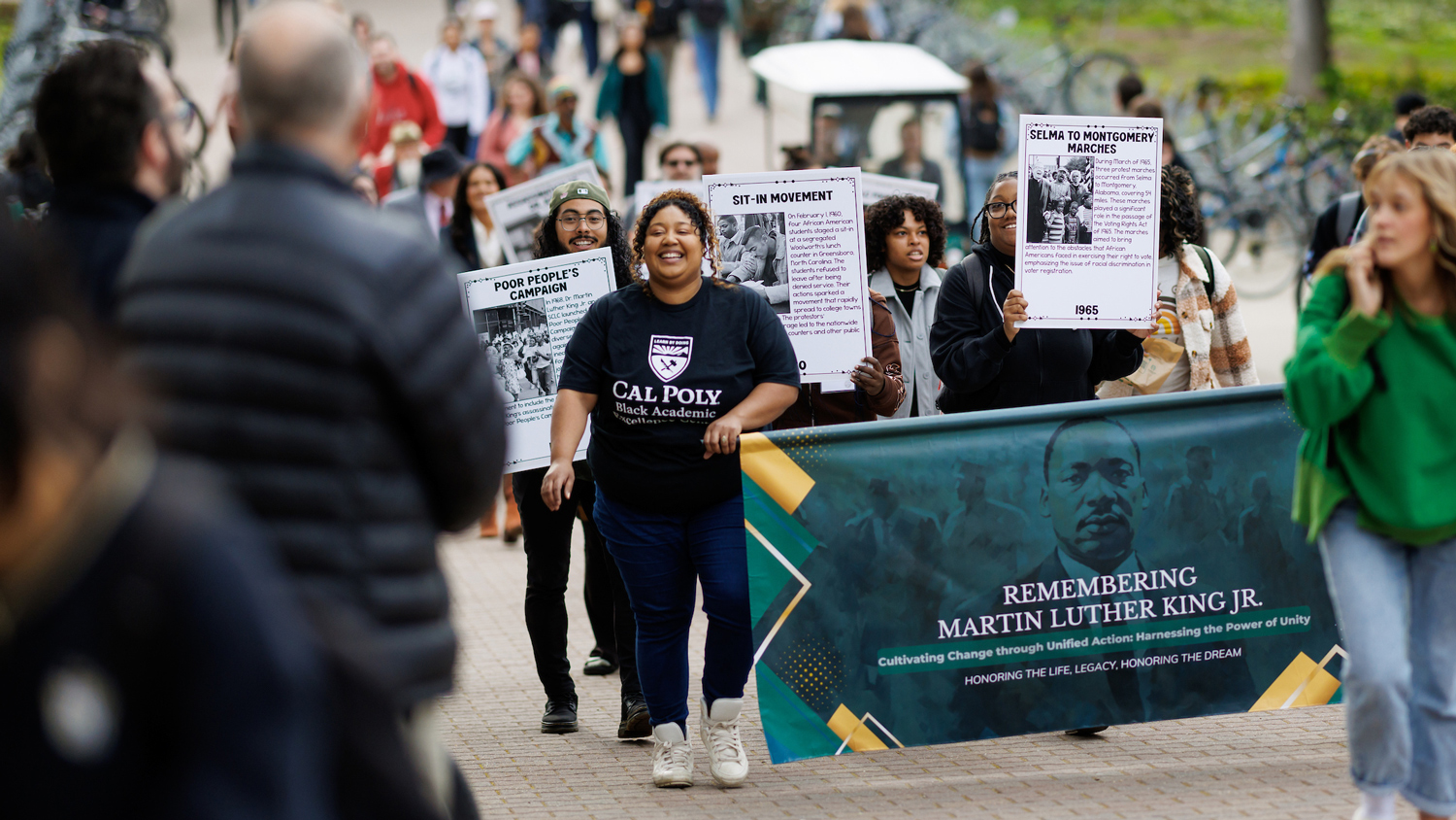 People march behind a green banner featuring a picture of Martin Luther King, Jr., holding signs depicting milestones in the Civil Rights Movement.