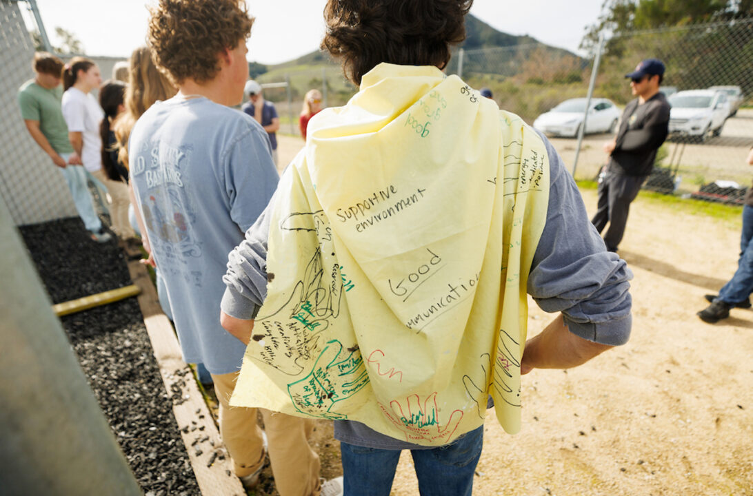 A person wears a yellow cape scrawled with hand-written words including supportive environment and good communication