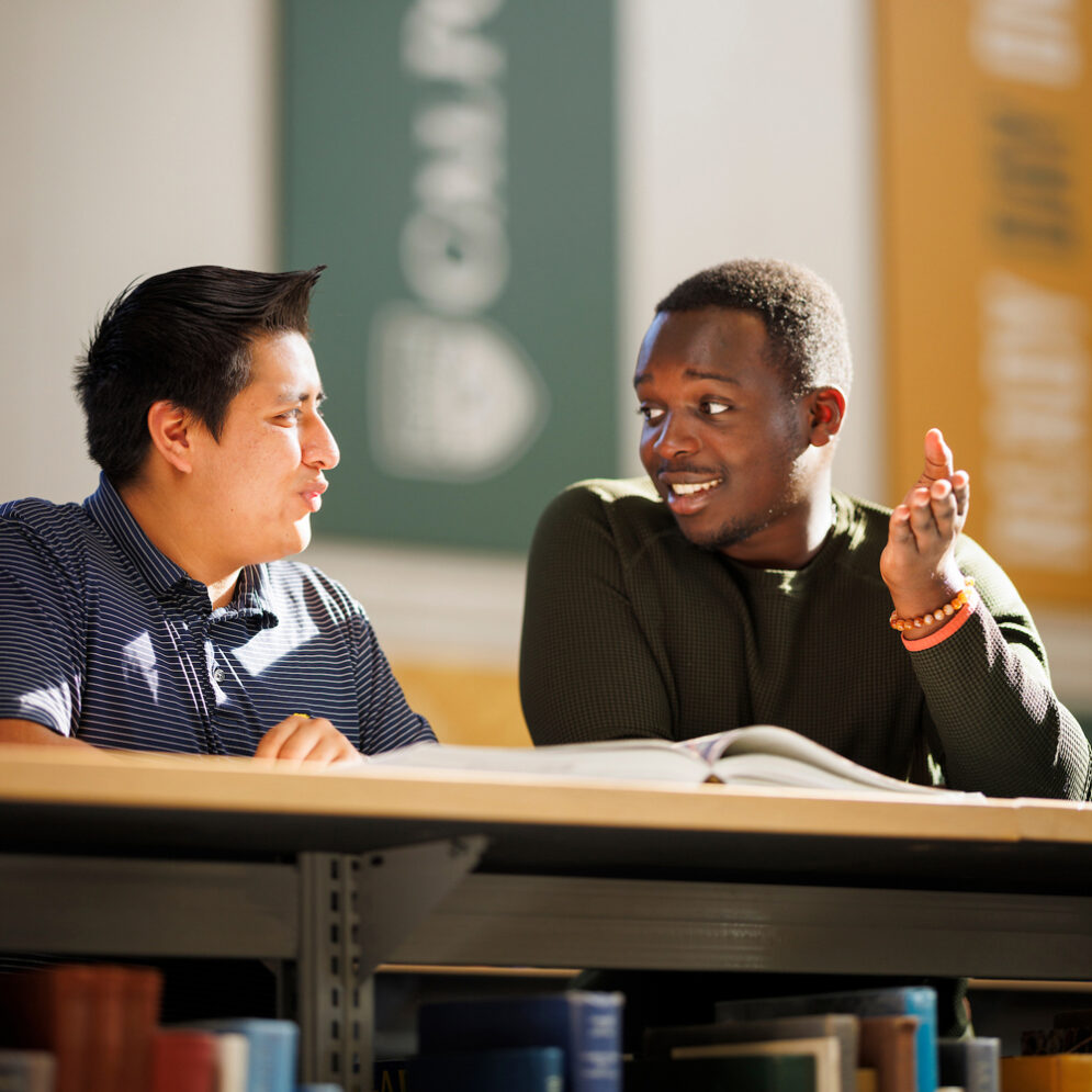 Two students discuss a book near a Cal Poly banner