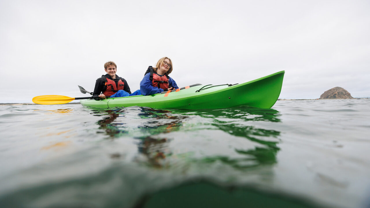 A young man and a girl ride in a green kayak on a bay.