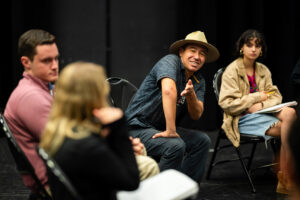 A man in a hat sitting in a chair gestures at a nearby student while discussing in a seated group.