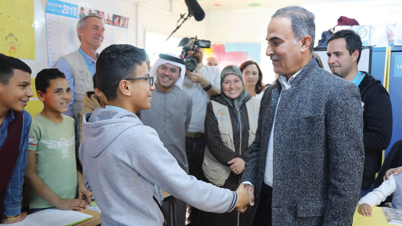 A man in a grey coat shakes hands with a boy in a sweater in front of a group of people