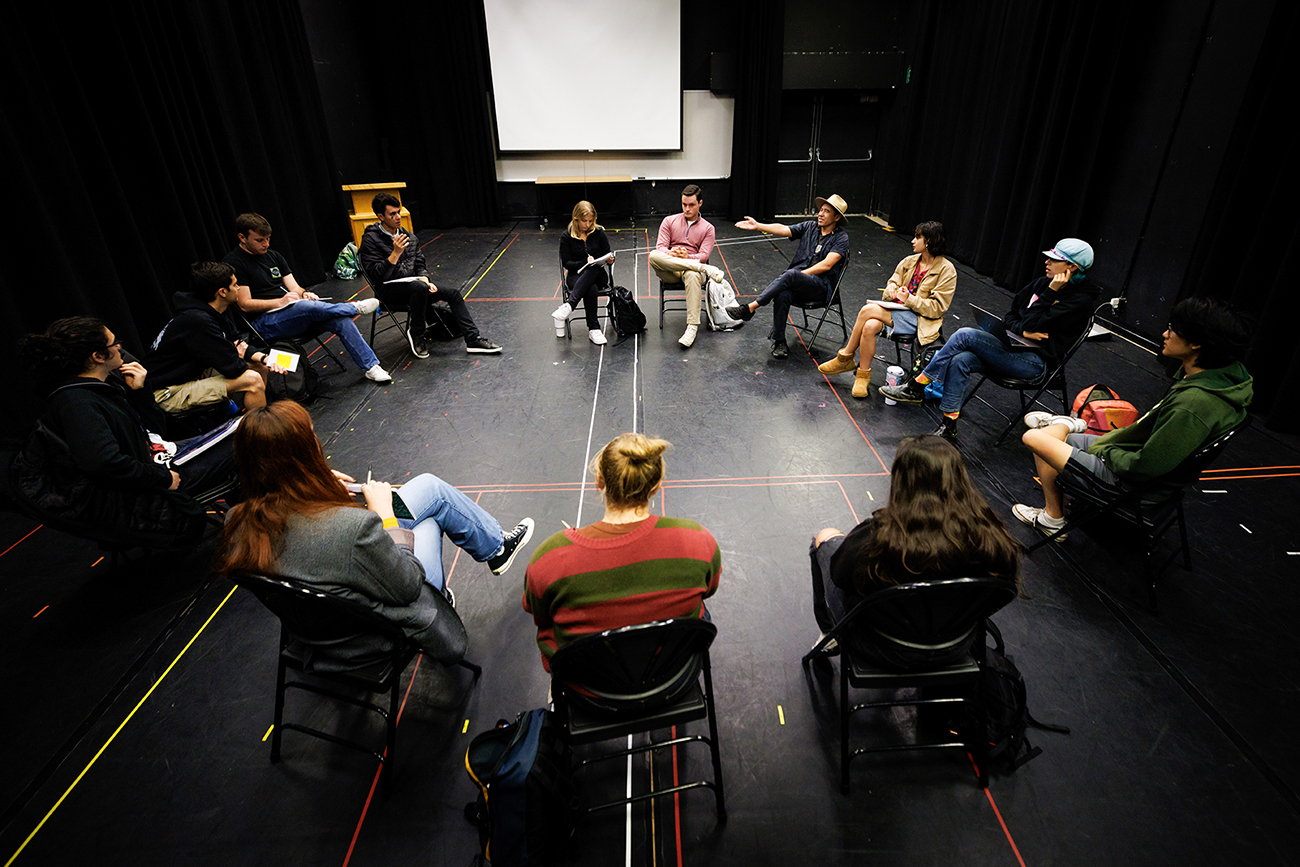 A group of students sits in a circle of chairs in an all black room.