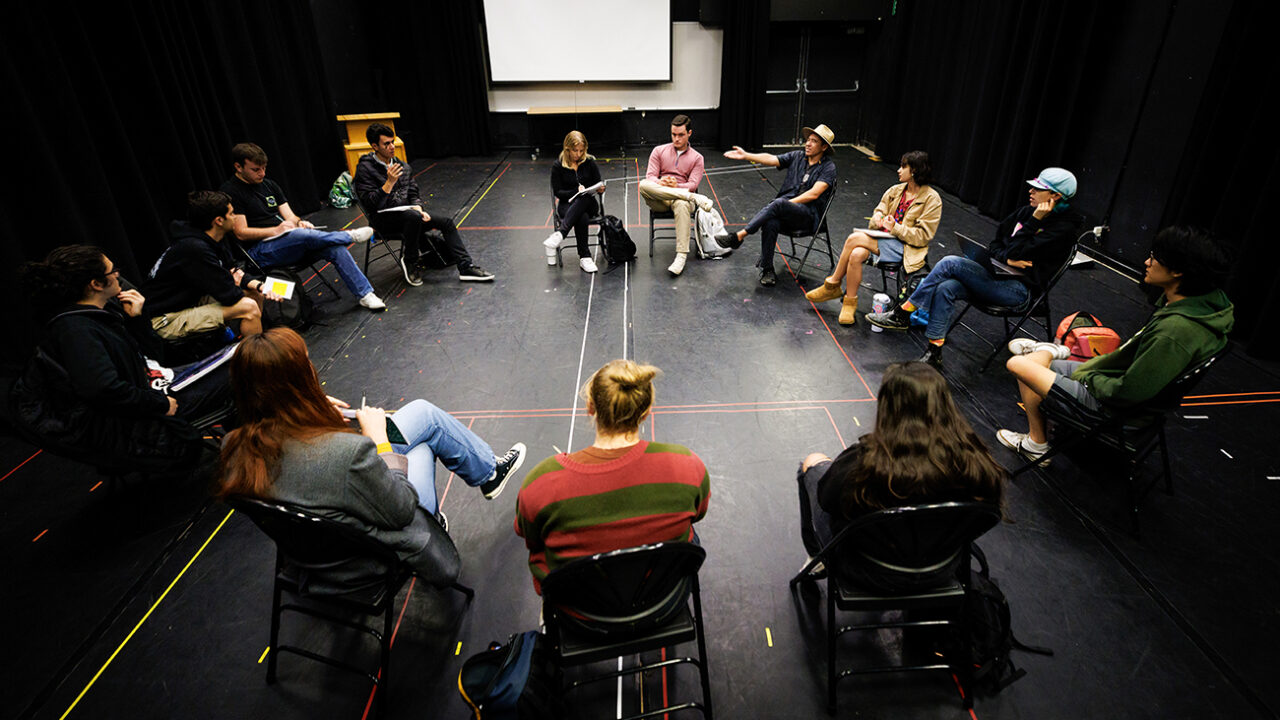 A group of students sits in a circle of chairs in an all black room.