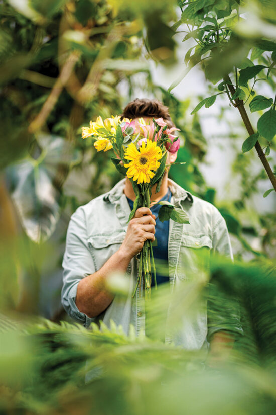 A man in a blue shirt stands among plants, with his face obscured by a bouquet of flowers