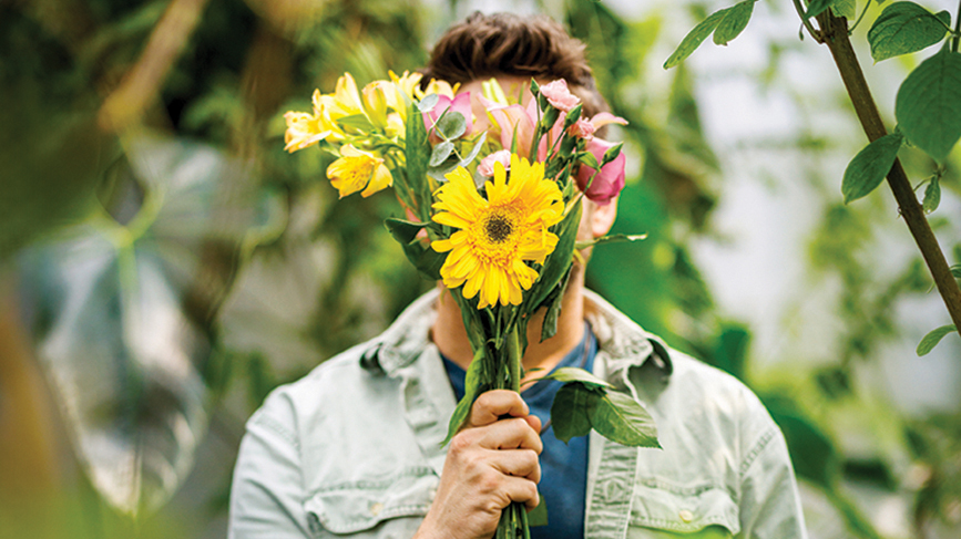 A man in a blue shirt stands among plants, with his face obscured by a bouquet of flowers