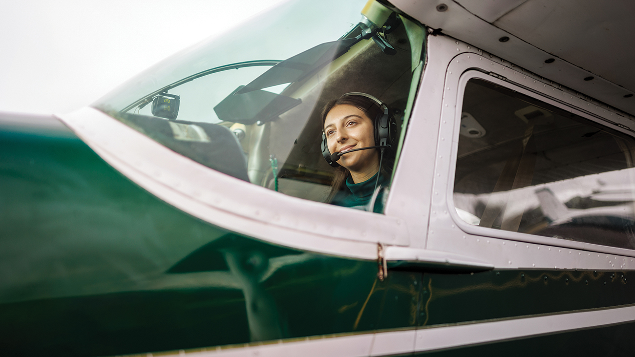 A young woman wearing a headset sits in the pilot seat of a green and white airplane