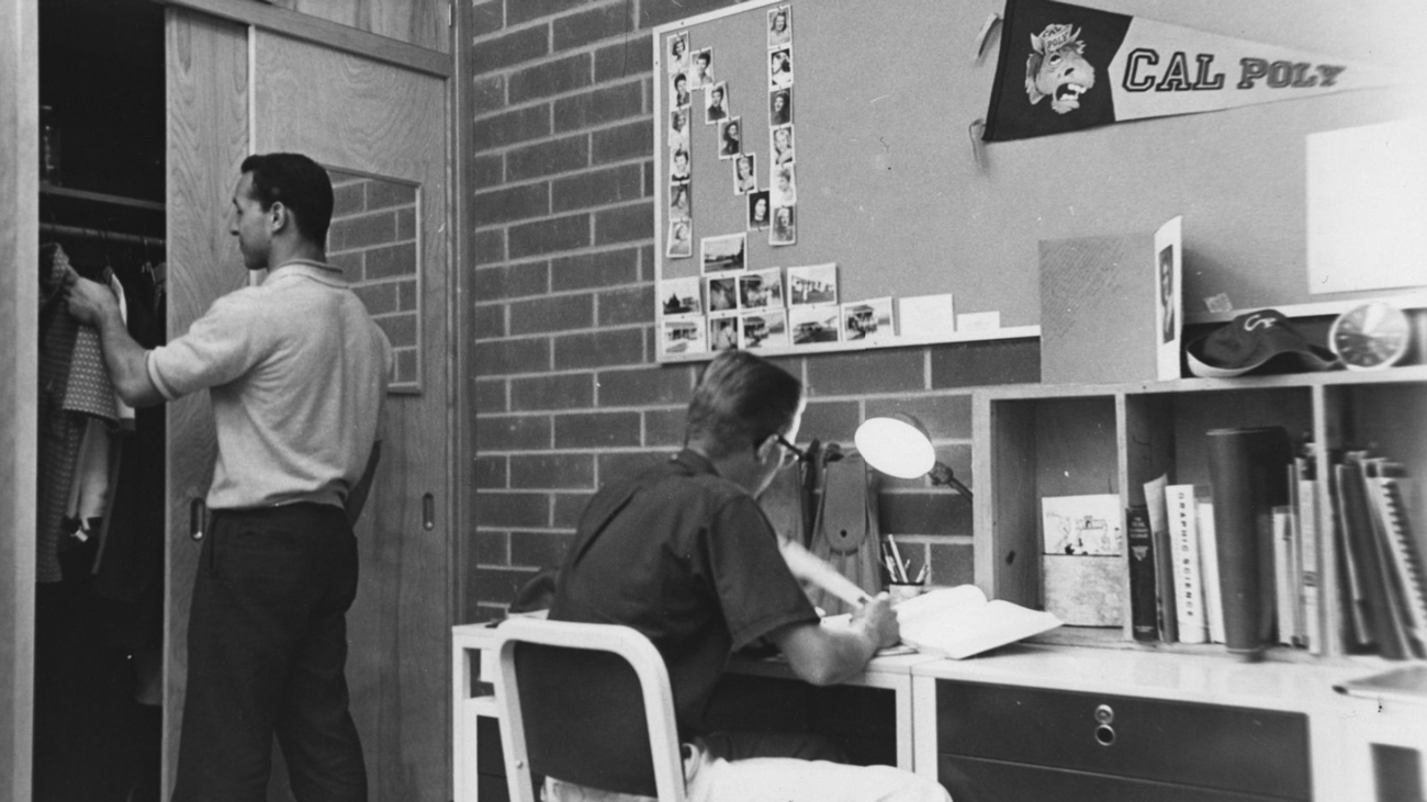 An archive black and white photo of two students in a dorm room with photos and a Cal Poly pennant hung on the cork board