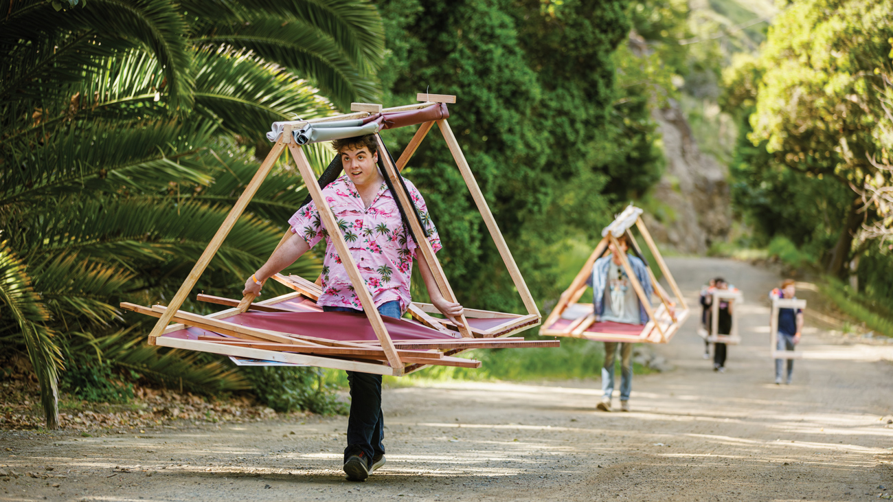 Four students carry wooden structures on a dirt road surrounded by greenery
