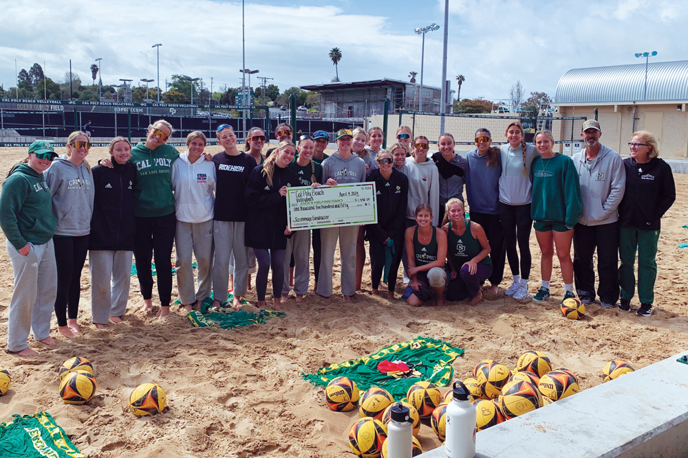 A group of Cal Poly beach volleyball players smile on the sand while holding a check for a charity organization