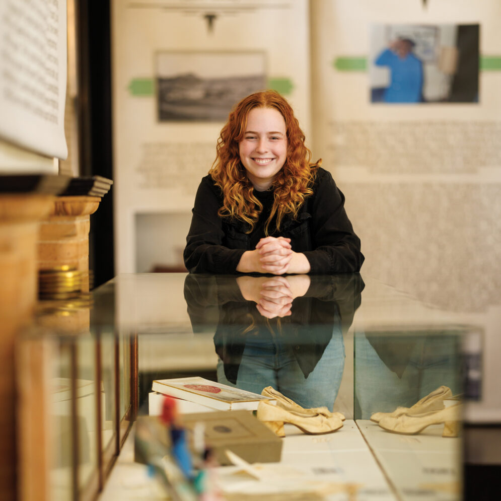 A student smiles with hands clasped near a museum exhibit with historical artifacts.