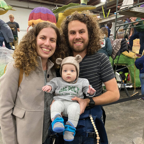 Two adults hold an infant wearing a Cal Poly sweater near a Cal Poly Rose Parade float under construction