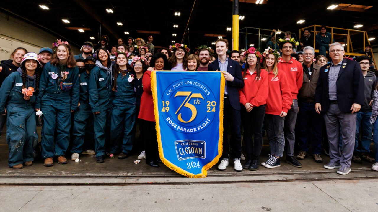 Students and campus leaders smile holding a blue banner celebrating the Rose Float program's 75th anniversary