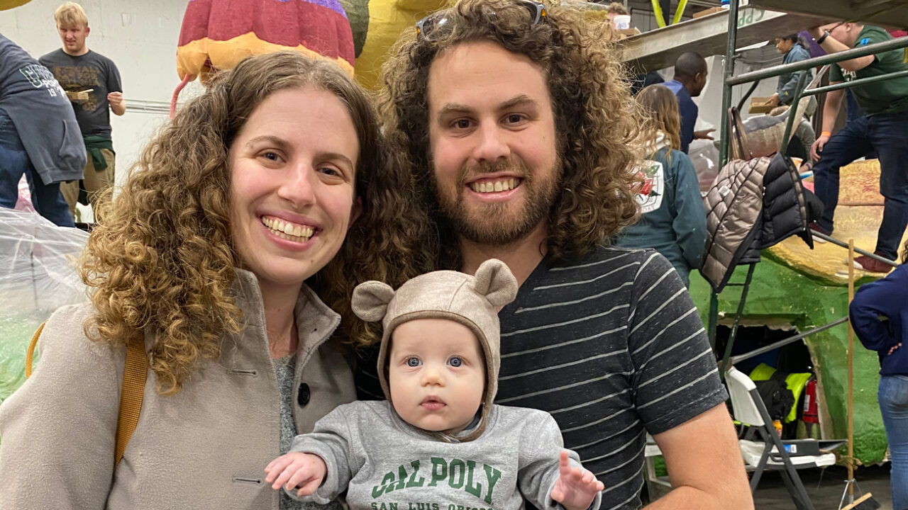 Two adults hold a baby wearing a Cal Poly sweater near a parade float under construction
