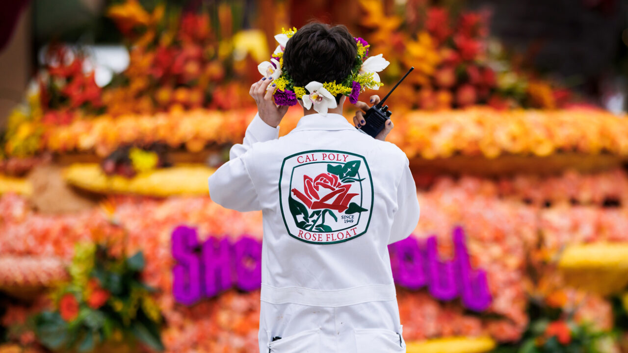 A Cal Poly student wearing a flower crown and a white jumpsuit with a Cal Poly Rose Float patch holds a walkie talkie and prepares to guide the Cal Poly Rose Float