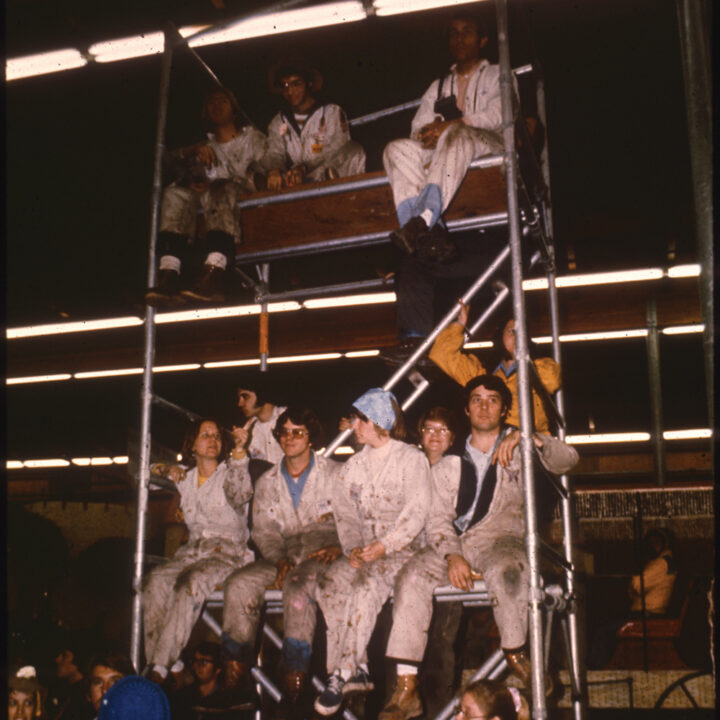 An archive image of students in coveralls sitting on scaffolding in a warehouse
