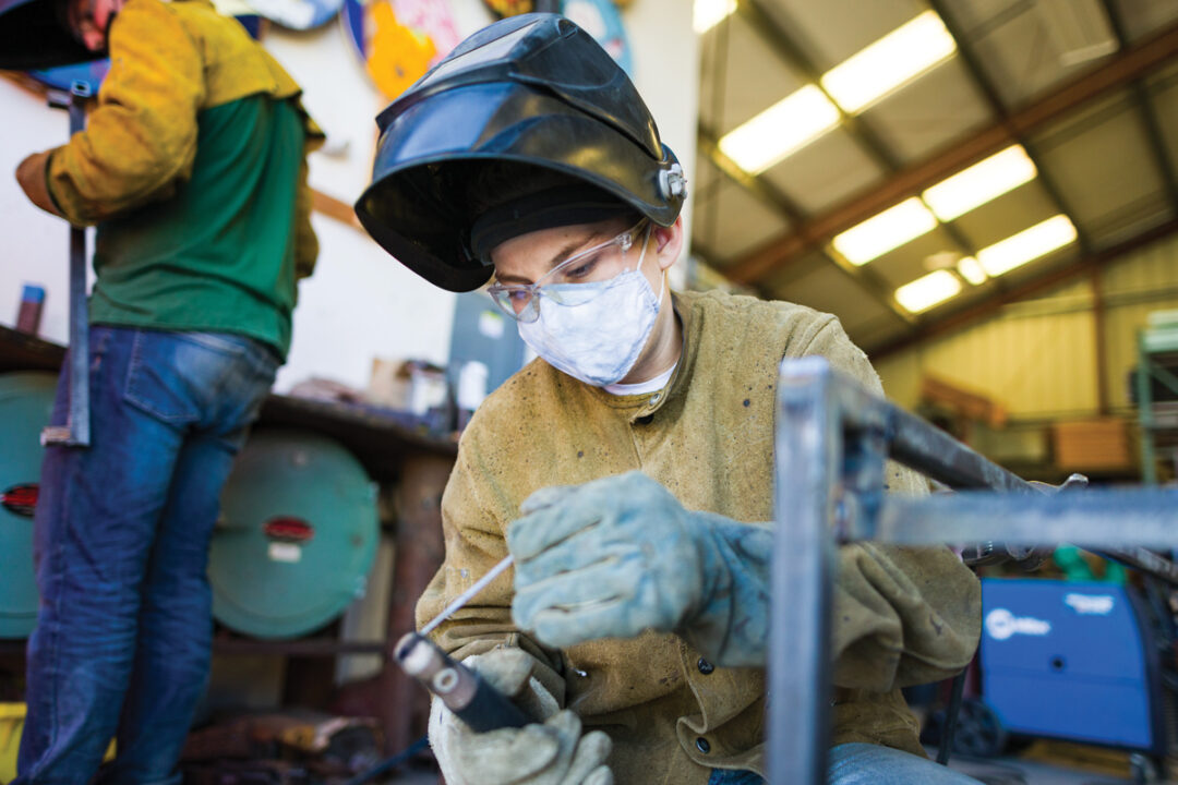 A welder wearing safety glasses, a welding helmet and leather apron prepares tools for fabrication.