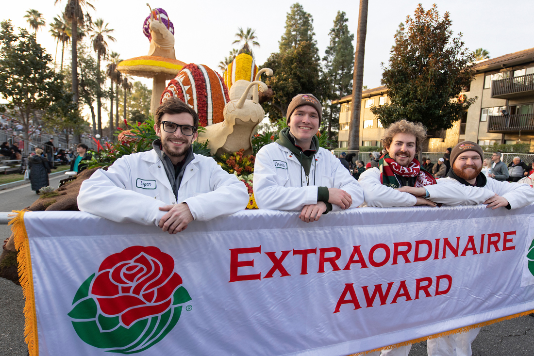 Four people in white coveralls stand next to a Rose Parade float and a banner that says Extraordinaire Award