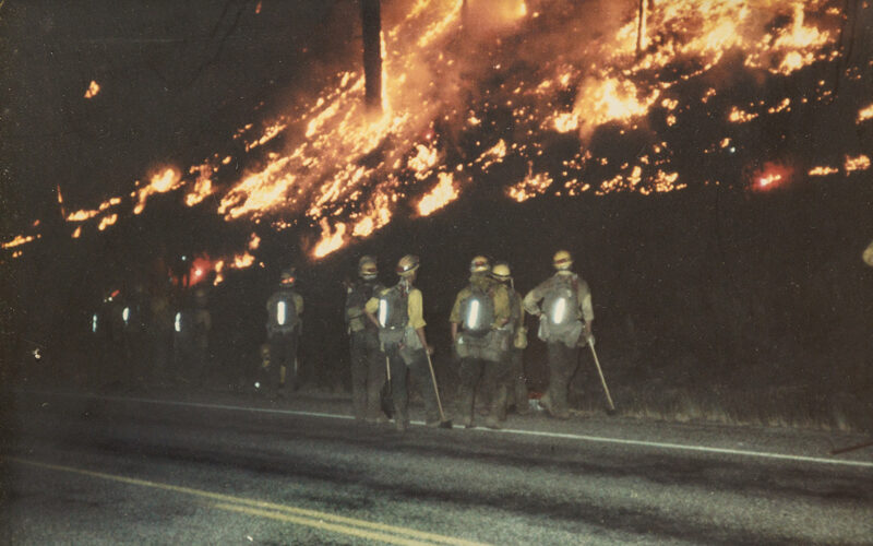 Firefighters standing on the side of a road with hand tools looking at a hillside on fire.