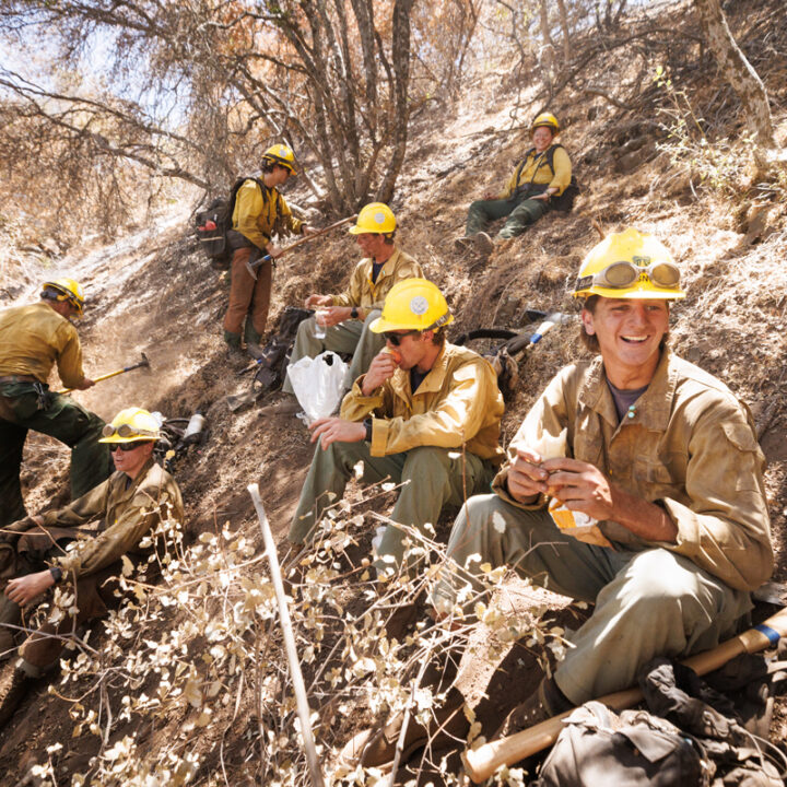 Smiling firefighters sit in hillside brush while taking a break and eating snacks.