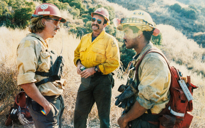 Three firefighters in yellow shirts and helmets talk on a hillside