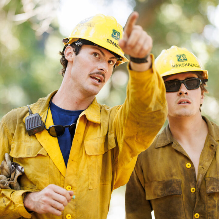 A firefighter wearing yellow jackets and helmets points forward while talking to a colleague