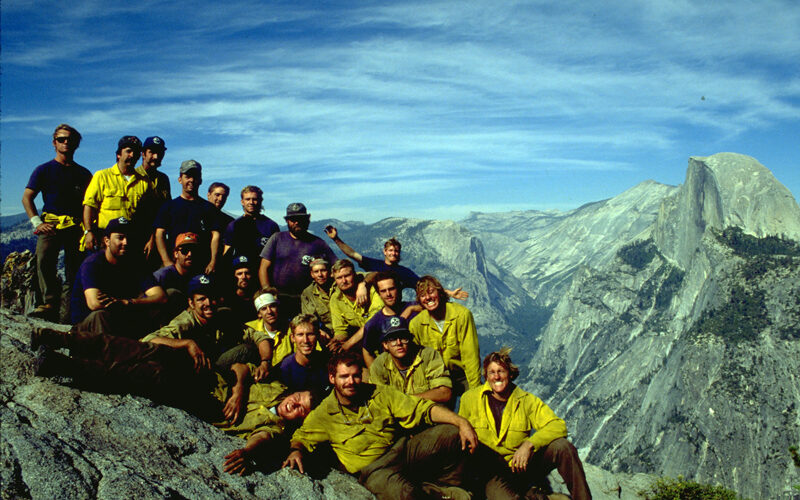 A crew of young firefighters smile at Glacier Point in Yosemite National Park with Half Dome in the distance.