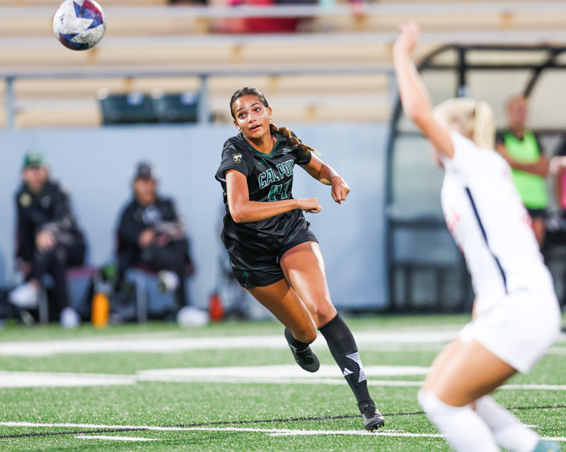 A women's soccer player runs toward a soccer ball in mid air next to a defender