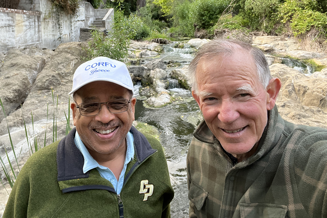 Two men in jackets smile for a selfie while hiking in a creek bed.