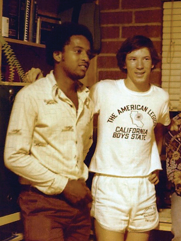 Two young men lean against a shelf in a brick dorm room in a vintage photo from the 70s.