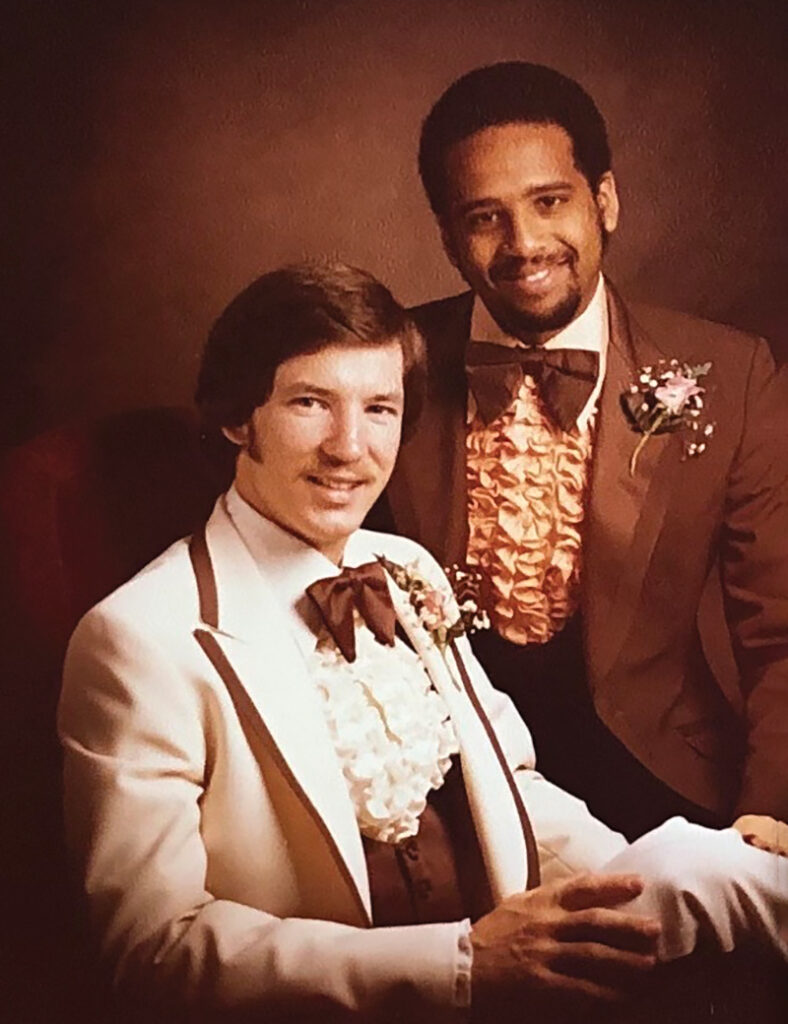 Two young men in frilly tuxedos smile for a sepia-toned portrait.