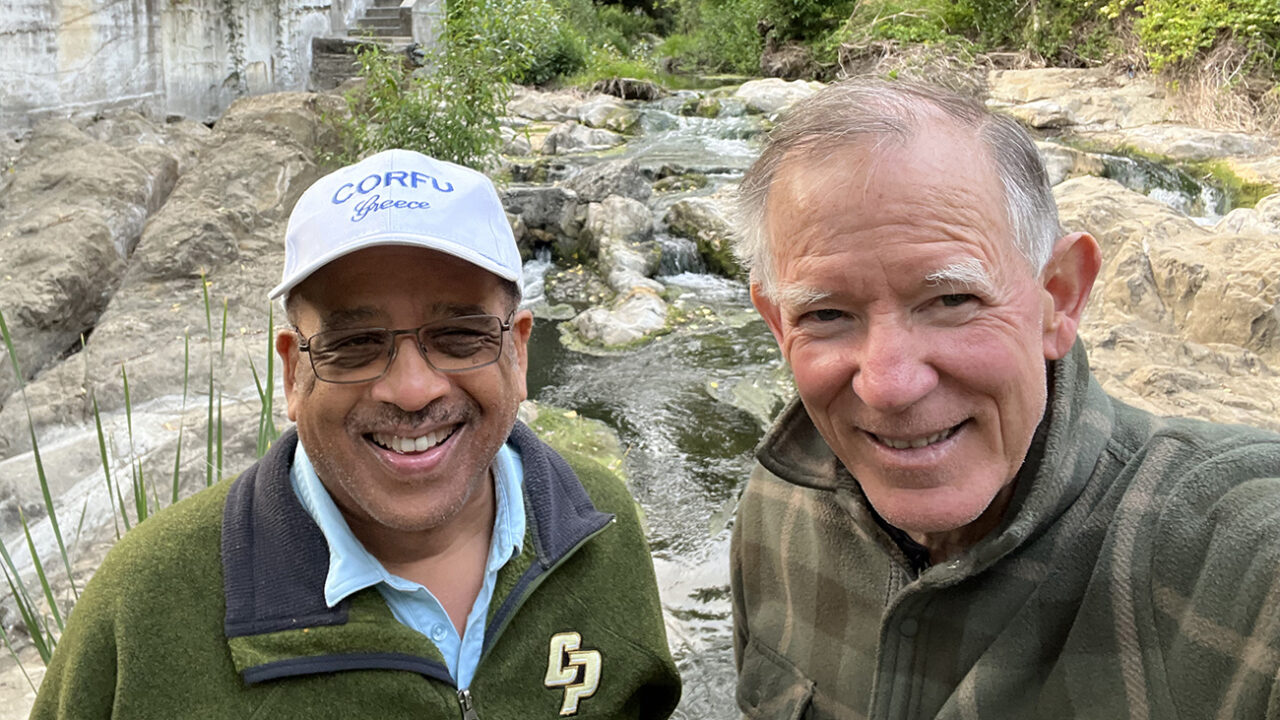 Two men in jackets smile for a selfie while hiking in a creek bed.