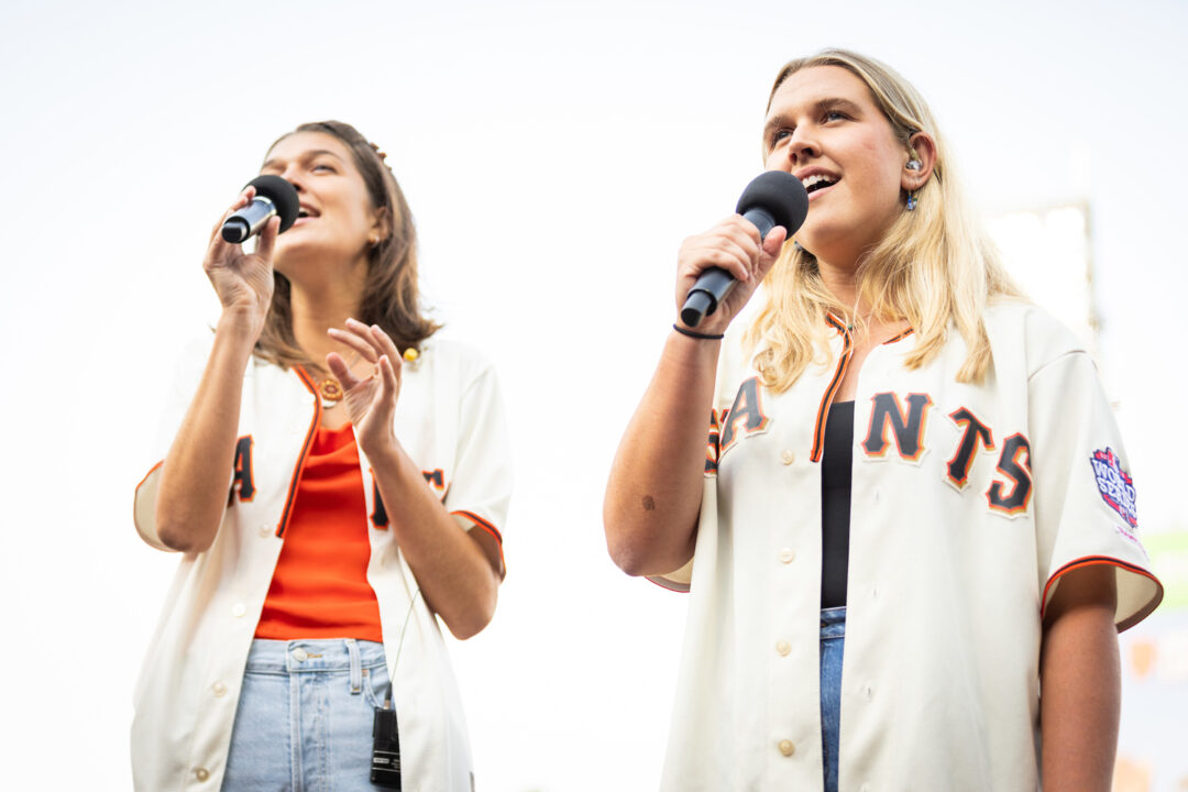 Two people wearing San Francisco Giants jerseys hold microphones as they sing the national anthem