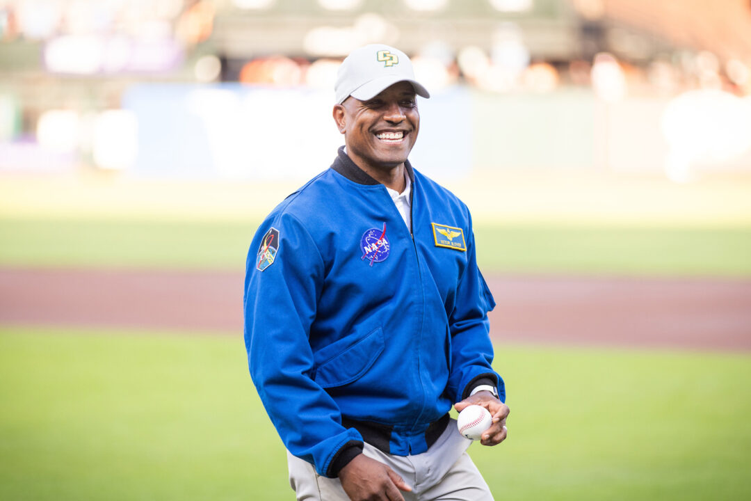 Astronaut Victor Glover wears a blue NASA jacket, a Cal Poly hat and holds a baseball