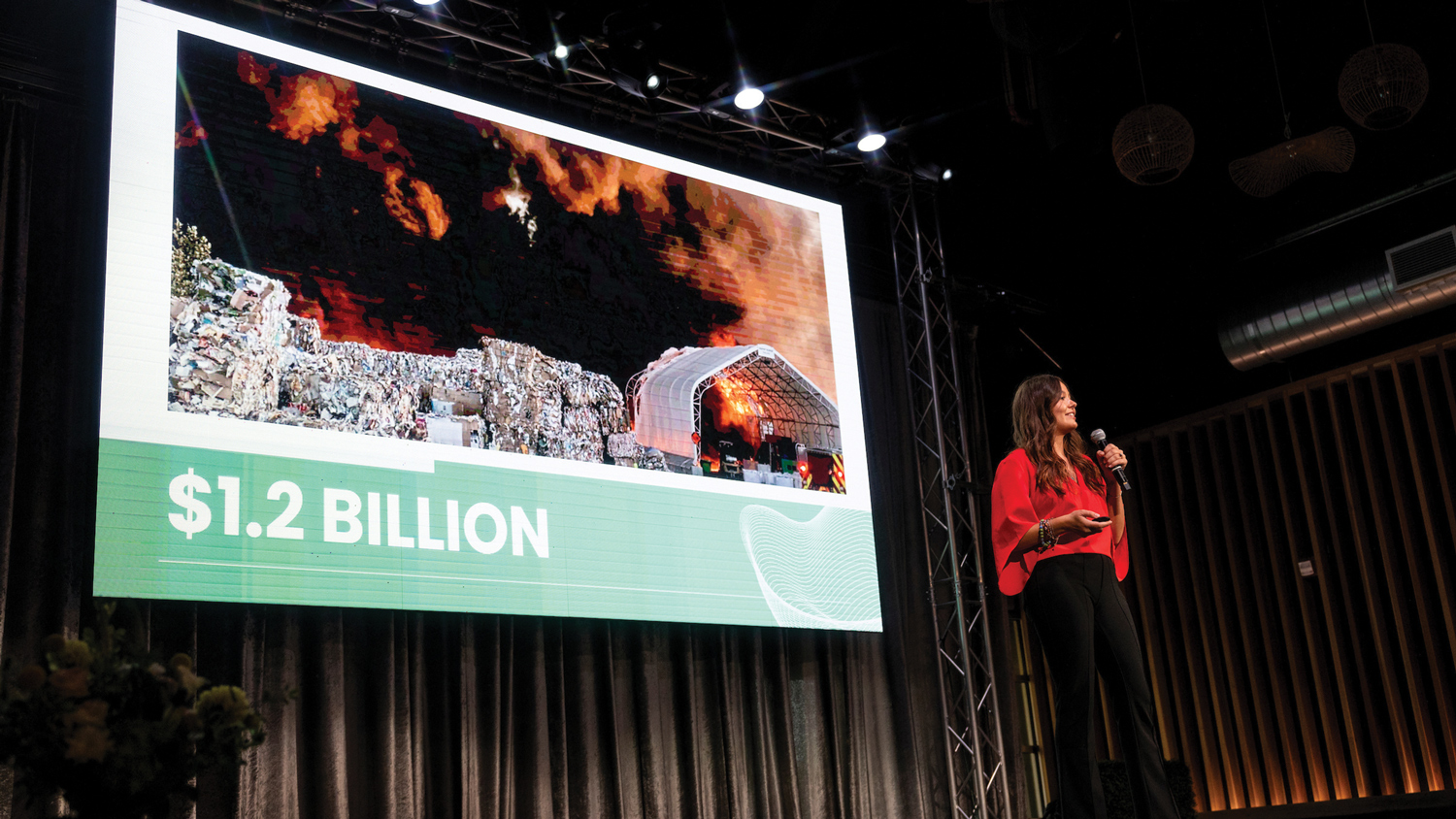 An entrepreneur in a red sweater stands on stage in front of a screen displaying a recycling center on fire with the number $1.2 billion