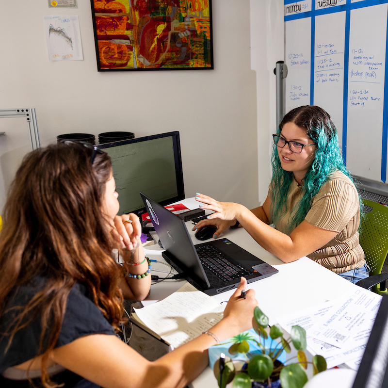 Two entrepreneurs sit at a desk near a whiteboard filled with notes