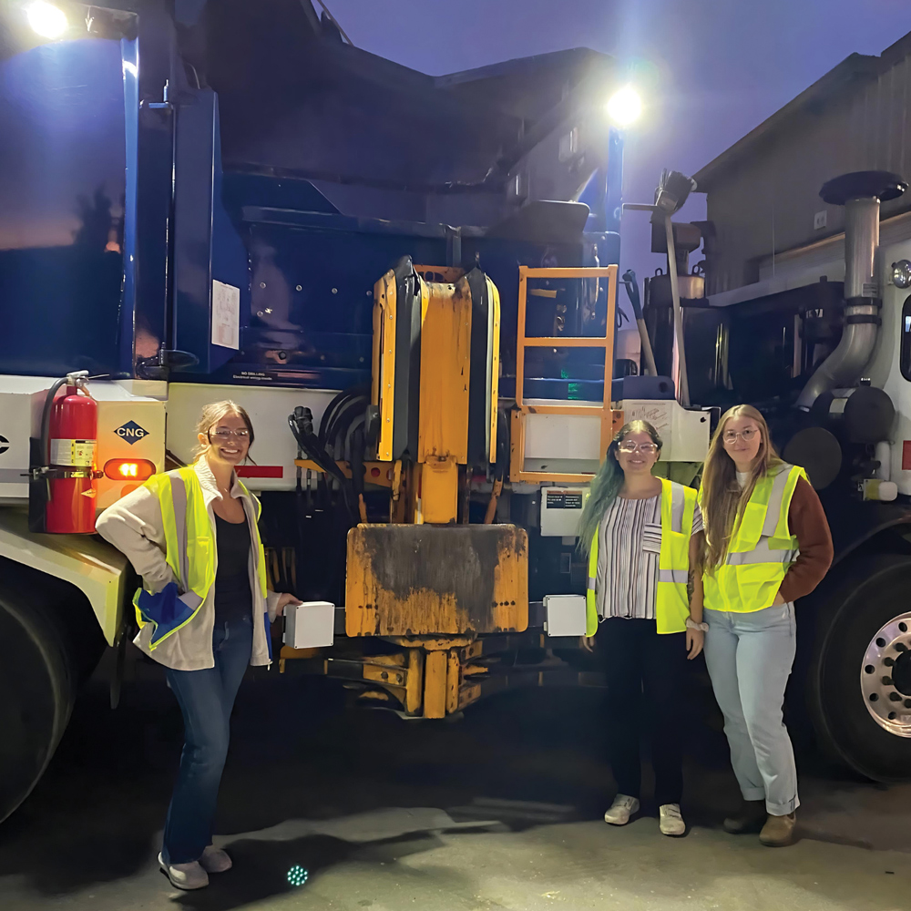 Three people in reflective vests stand next to a side-loading garbage truck