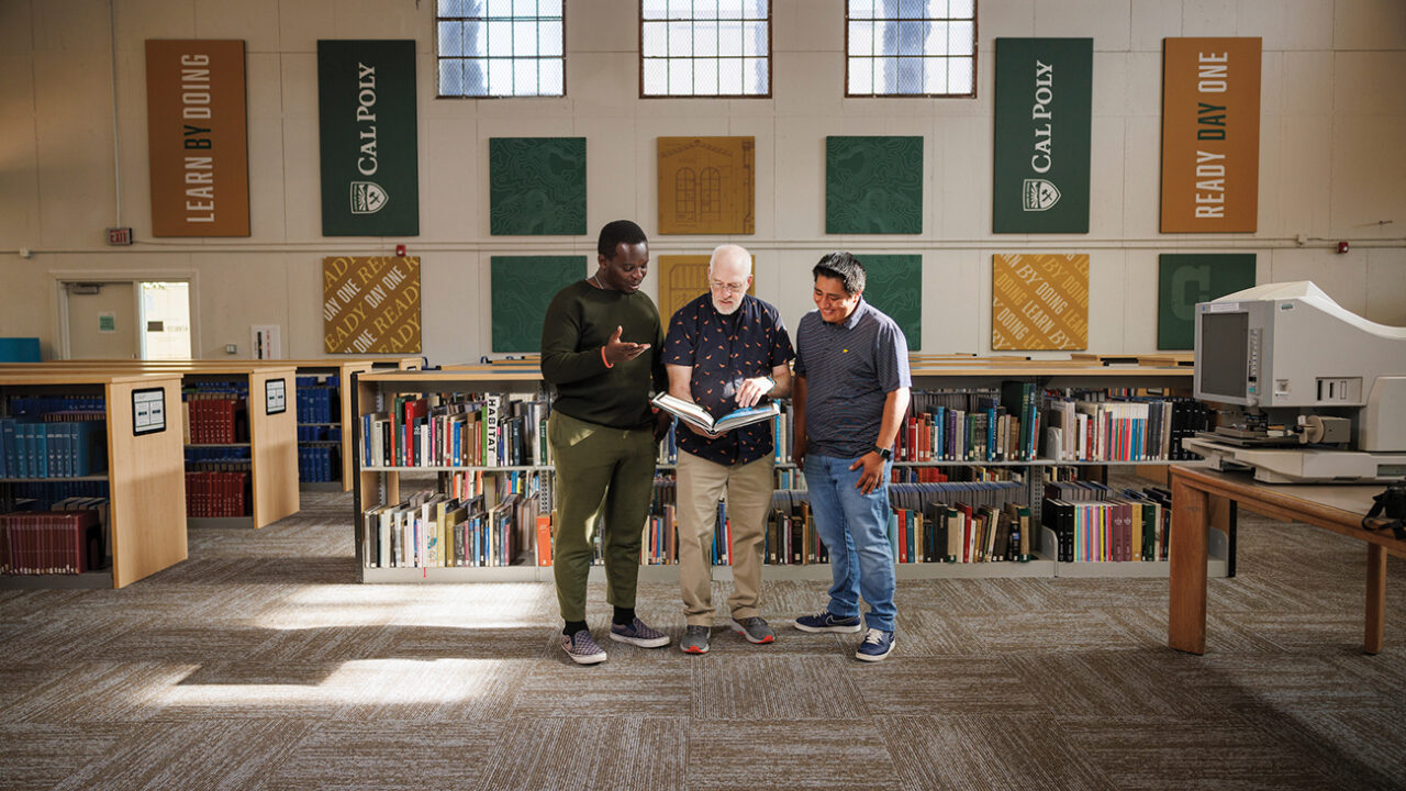 A librarian shows a large book to two students in a library with high windows and green and gold banners on its walls.