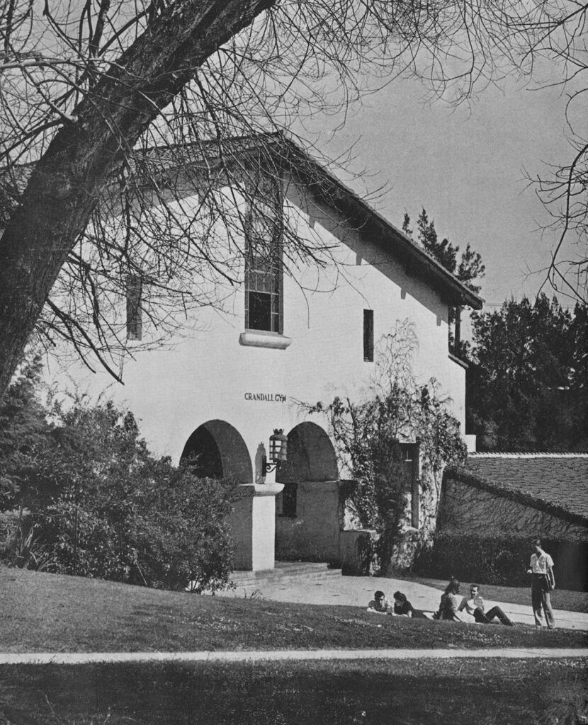 A black and white vintage photo of a tall mission-style building with students lounging on the lawn in front.