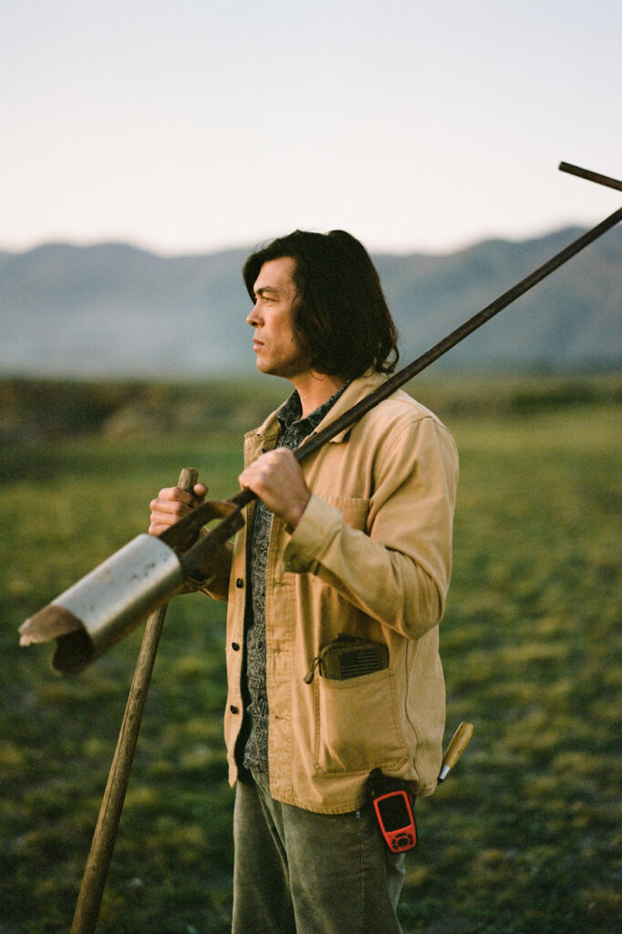 Jackson, in a jacket, stands on a grassy plain holding a shovel and a core sampling tool, with hills in the background.