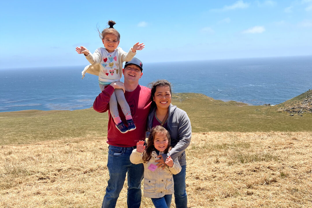 Two parents and two children smile on a hillside near the Pacific Ocean