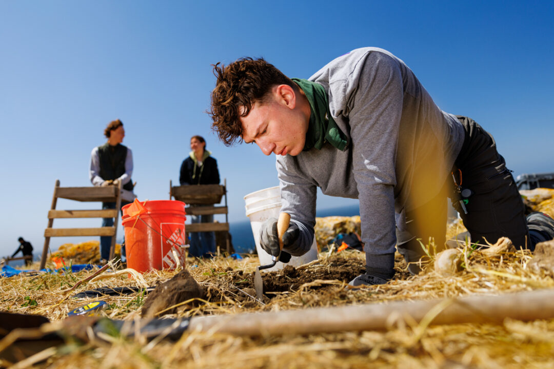 A student on his hands and knees uses a trowel to dig through dirt near an orange bucket and two students using shaker screens.