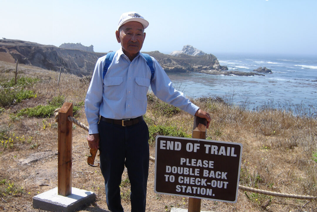 A man in a hat stands next to a coastal trail that says End of Trail Please Double Back to Check-Out Station