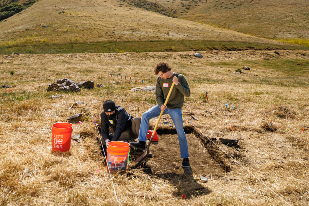 A person standing in a square excavation trench uses a shovel while another kneels in the dirt near two orange buckets