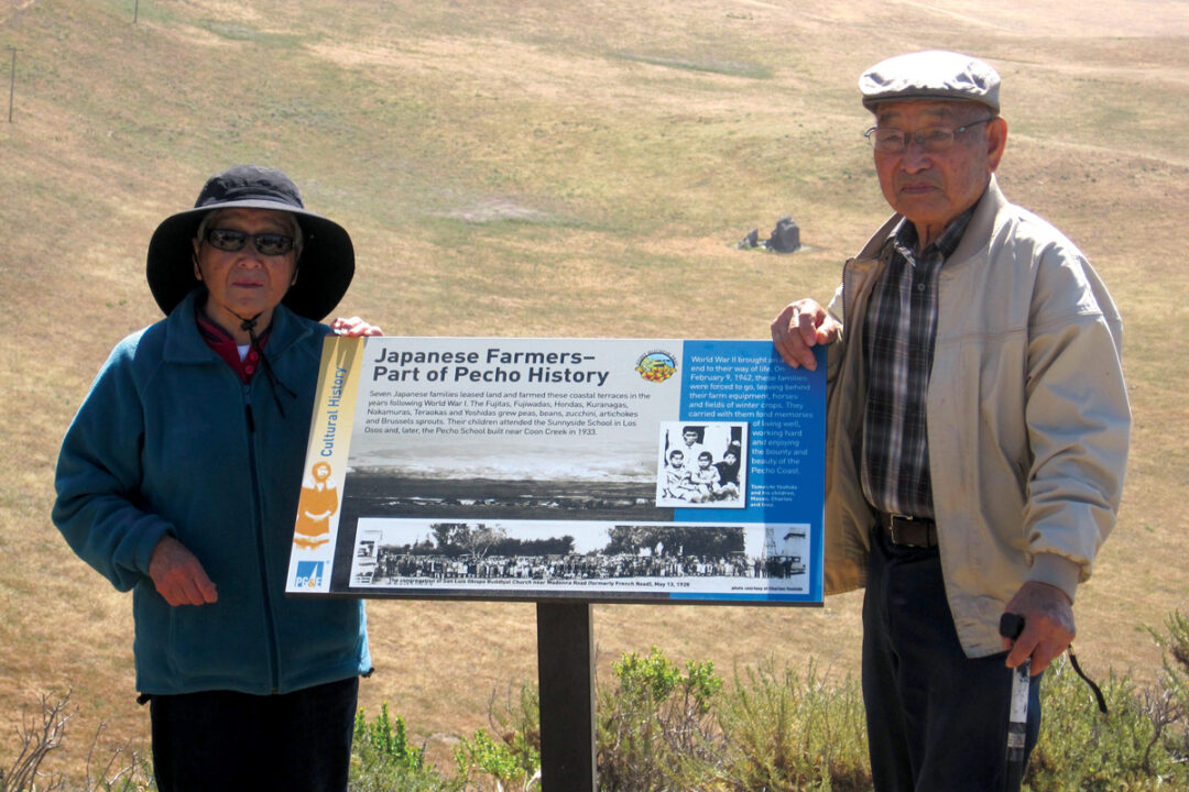 Two elderly people stand next to a trail marker titled Japanese Farmers — Part of Pecho History