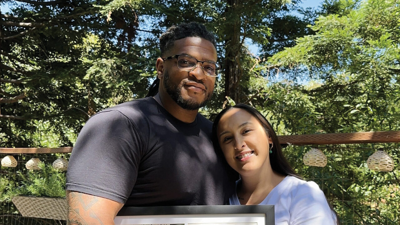 A husband and wife smile together under a green tree