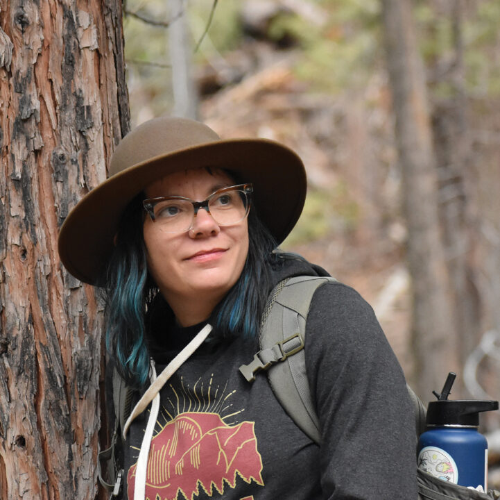 A woman in glasses and a wide brim hat leans against a tree in the forest.