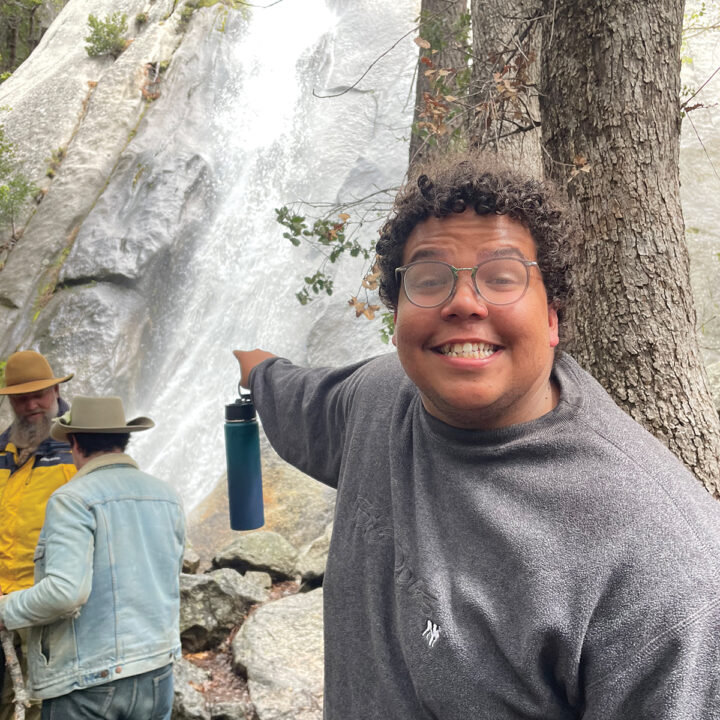 A young man in damp glasses smiles and points at a waterfall.