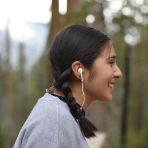 A young woman smiling and wearing earbuds stands among trees.