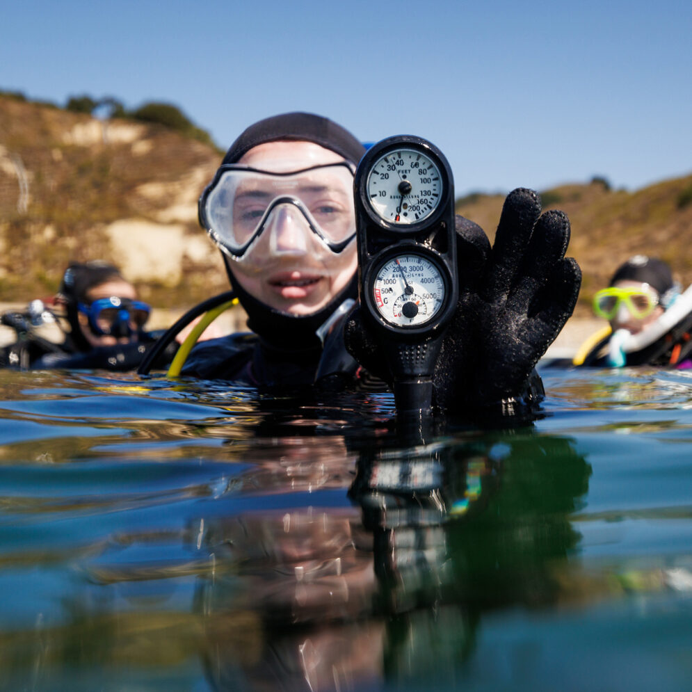 A student scuba diver holds a pressure gauge at the surface of the ocean near two other scuba divers